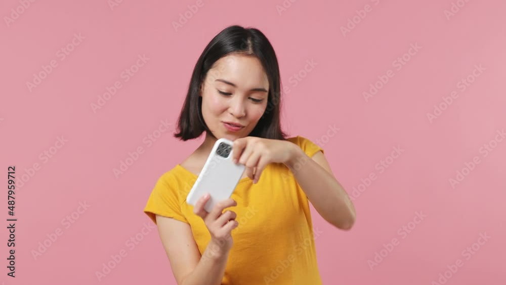 Gambling young woman of Asian ethnicity 20s wears yellow t-shirt using play racing on mobile cell phone hold gadget for pc video games isolated on plain pastel light pink background studio portrait