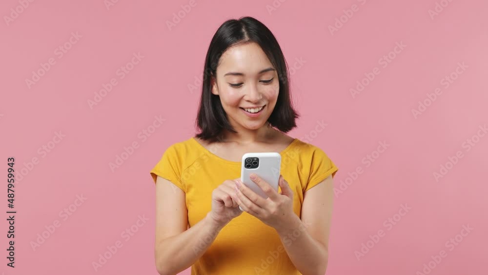 Dreamful minded smart young woman of Asian ethnicity 20s wears yellow t-shirt hold using mobile cell phone swipe typing browsing chatting isolated on plain pastel light pink background studio portrait Dreamful minded smart young woman of Asian ethnicity 20s wears yellow t-shirt hold using mobile cell phone swipe typing browsing chatting isolated on plain pastel light pink background studio portrait