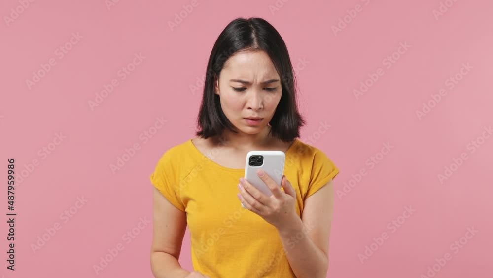 Sad young woman of Asian ethnicity 20s wears yellow t-shirt using mobile cell phone hears fake news unexpected rumor has some problems isolated on plain pastel light pink background studio portrait