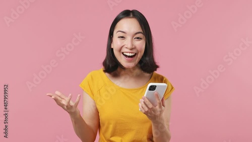 Surprised joyful young woman of Asian ethnicity 20s in yellow t-shirt hold use mobile cell phone typing say wow yes doing winner gesture isolated on plain pastel light pink background studio portrait photo