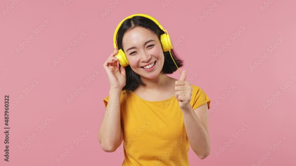 Vivid cheerful young woman of Asian ethnicity 20s wears yellow t-shirt listen music in headphones dance sing song have fun enjoy relax isolated on plain pastel light pink background studio portrait