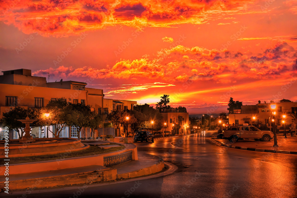 Fototapeta premium Inusual escena nocturna de una avenida en La Marsa (Túnez) sin vehículos ni personas en un atardecer con un cielo rojo que tiñe toda la avenida con su color predominante.