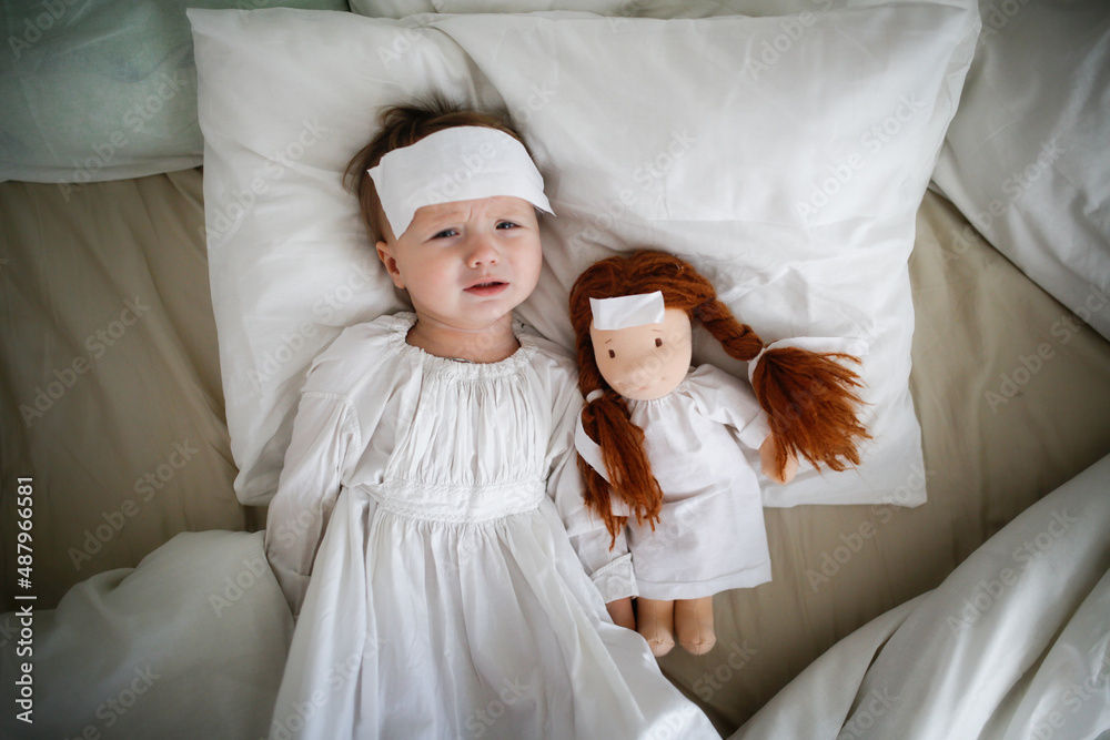 Cute baby with a doll with a compress on his head, a sick child in bed, bed rest during illness. Comfort and play in bed