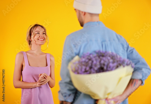 Young man hiding flowers behind his back. Womans day, spring selebration , 8 March concept. Beautiful woman in purple dree on yellow background. photo