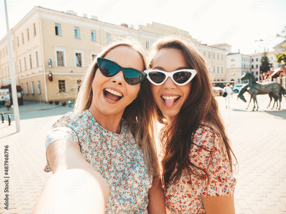 Two young beautiful smiling hipster female in trendy summer dress clothes.Sexy carefree women posing on the street background. Positive models giving air kiss, taking Pov selfie
