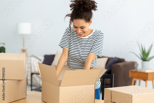 Young smiling African American woman unpacking boxes standing at home. Unpacking parcel, delivery, moving to new house concept. Female Student moves to new home