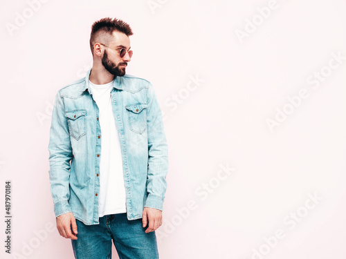 Portrait of handsome confident stylish hipster lambersexual model.Man dressed in jacket and jeans. Fashion male posing near wall in studio in sunglasses