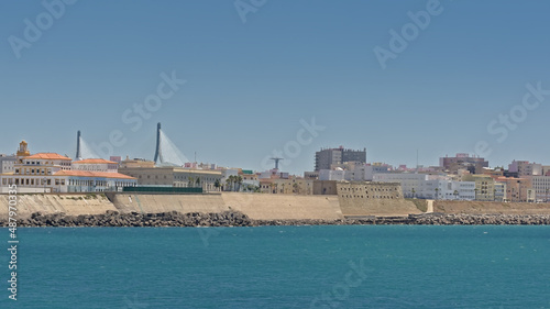 Coast of the deep blue atlantic ocean in Cadiz, with historic fortification, apartment buildings and pylons of a modern bridge. Andalusia Spain 