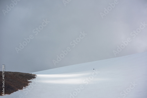 Awesome scenery with small man on large snowy mountain in sunlight under gray cloudy sky. Atmospheric landscape with sunshine on high snow mountain at changeable weather. Sunlit mountain in overcast.