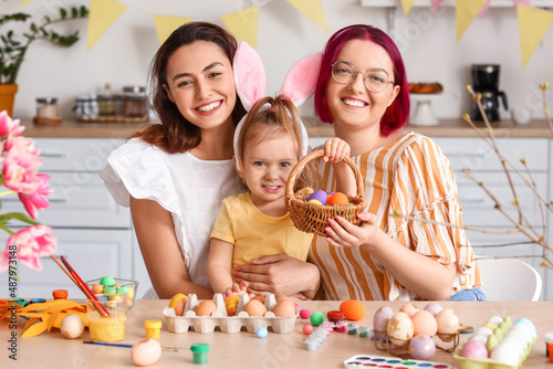 Young lesbian couple with little daughter during painting of Easter eggs in kitchen