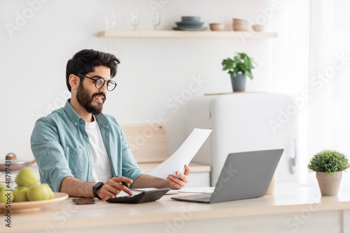 Savings and finances concept. Young arab guy using calculator and laptop computer, calculating taxes