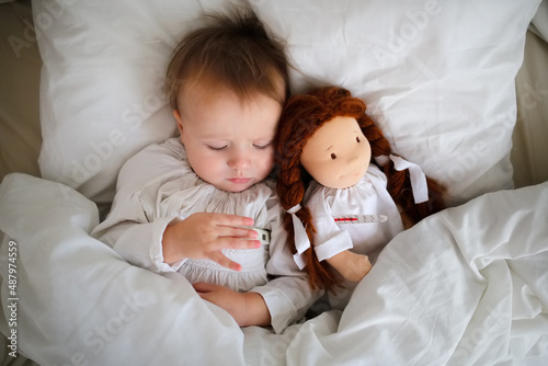 A cute Caucasian baby with a doll and with a thermometer, a sick child lies in bed with a toy, bed rest during illness. Comfort and play in bed