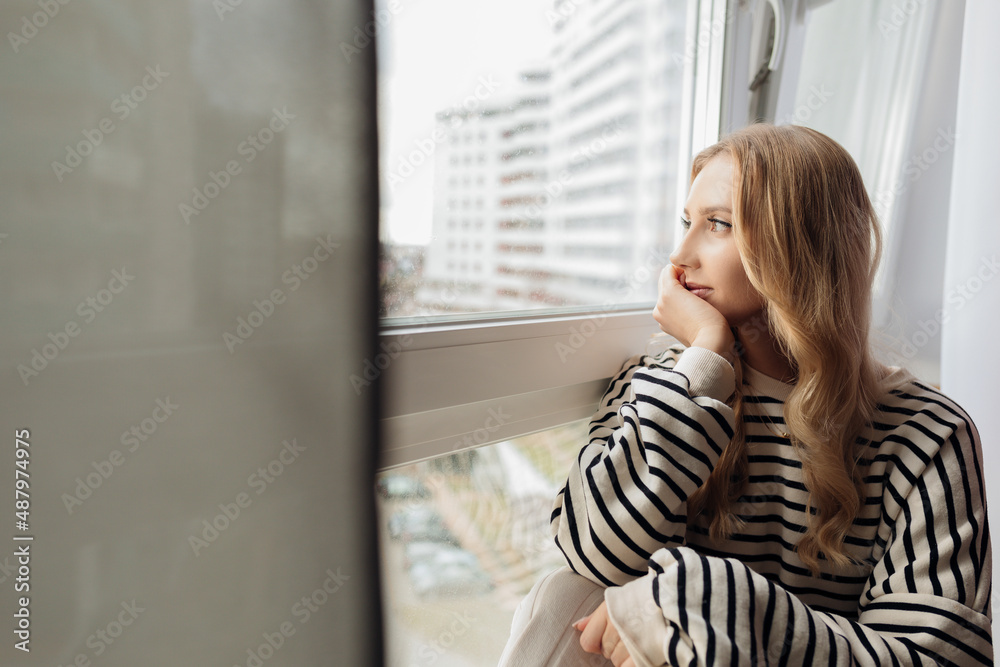 Portrait of pensive thoughtful blond pretty dreaming depressed woman sitting window sill looking distance street. Side