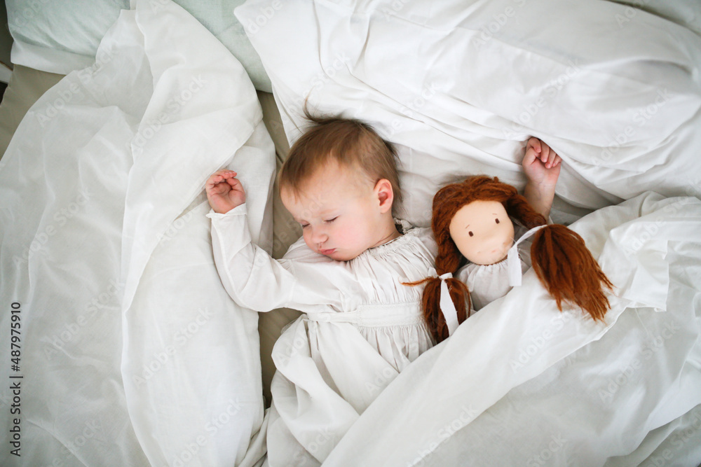 A cute Caucasian baby with a doll sleeping in bed , a sick child lies in bed with a toy, bed rest during illness. Comfort and play in bedroom