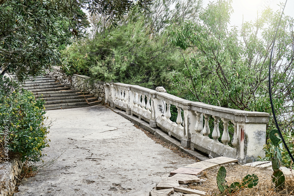Fototapeta premium Stone staircase and shabby balcony with figure railing near garden. Branches of trees leaning on stairs and concrete floor at bright sunlight
