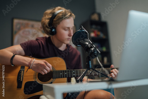 Home sound studio young teenager portrait playing guitar in Headphones with the microphone on the foreground. Modern audio recording technology concept image.