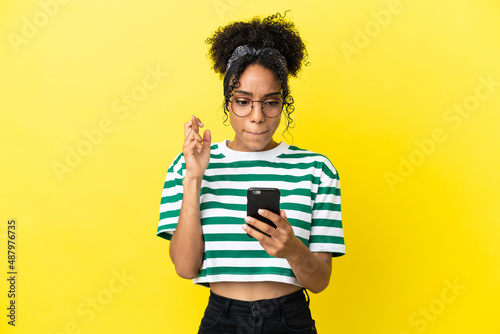 Young african american woman isolated on yellow background using mobile phone with fingers crossing