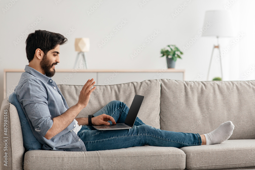Online communication concept. Arab man making virtual video call on laptop, talking and waving hand at webcam Online communication concept. Arab man making virtual video call on laptop, talking and waving hand at webcam