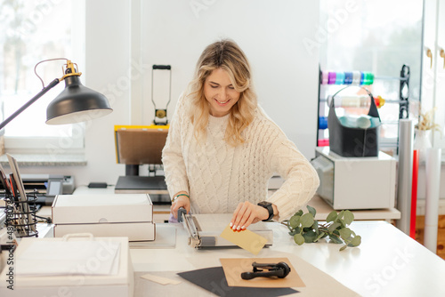 Professional smiling blond woman in sweater worker using printer cutter in workshop. Print office. Carving sheets paper  photo