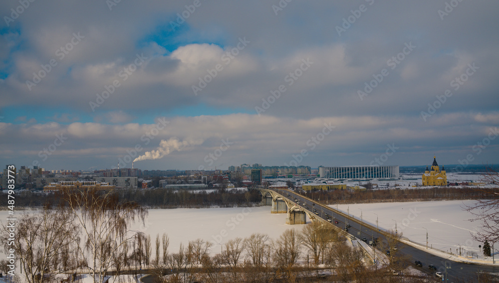 An urban landscape with a frozen and snow-covered river on the background of a big city. A road bridge, a modern stadium and an ancient cathedral. The spring blue sky emerging through the clouds.