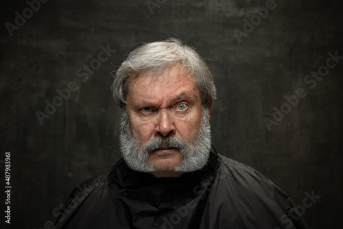 Dramatic portrait of emotional senior bearded man getting beard grooming isolated on dark vintage background. Concept of emotions, fashion, beauty, self-reinvention