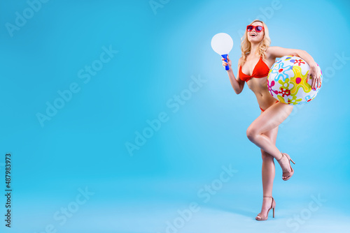 Young sunny beautiful woman in red swimsuit, glasses ready to play beach games. Blond smiling girl is holding matkot racket and colorful ball against blue background. Summer mood. Sea vacation concept