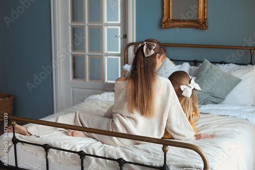 Portrait from behind of loving beautiful young mother and little daughter with long gorgeous hair with a trendy hairpin bow at home on bedroom sitting in cozy bed. Happy mothers day photo