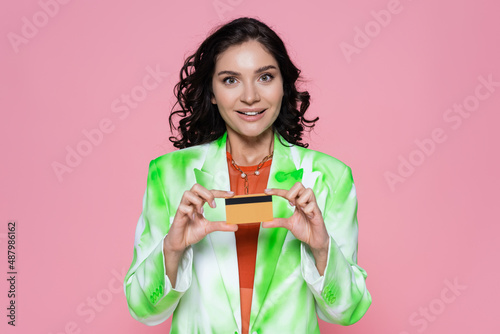 cheerful young woman in tie-dye blazer holding credit card isolated on pink.