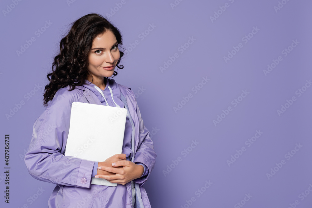 cheerful young woman in violet raincoat holding laptop isolated on purple. cheerful young woman in violet raincoat holding laptop isolated on purple.