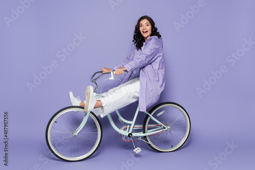 Fototapeta Naklejka Na Ścianę i Meble -  full length of amazed young woman in violet raincoat riding bicycle on purple.