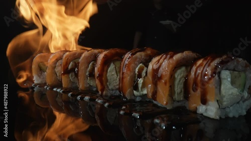 Philadelphia sushi roll with salmon and unagi sauce roasted on fire. Fire envelops set of sushi on black background close up. Sushi roll grill with salmon, rice, avocado and cheese. Slow motion. photo