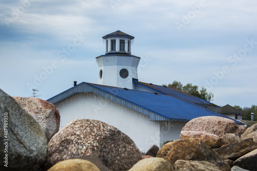 a white building with a blue roof and a small lighthouse tower on beach through large stones