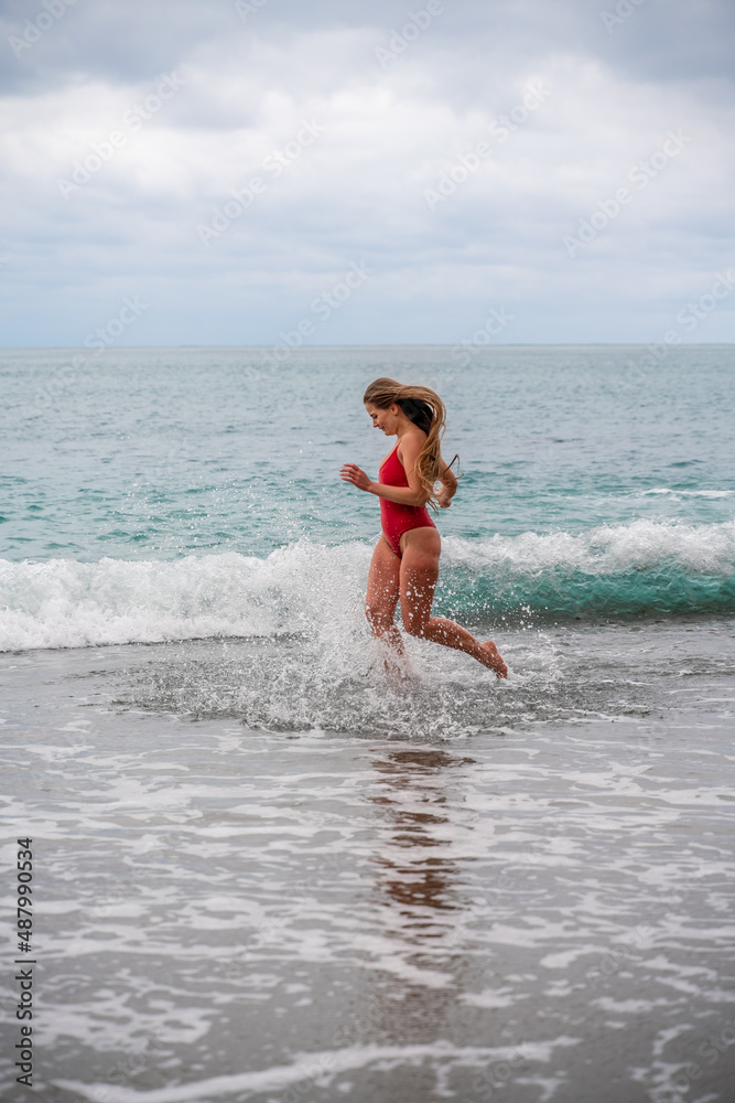 A beautiful and sexy brunette in a red swimsuit on a pebble beach, Running along the shore in the foam of the waves A beautiful and sexy brunette in a red swimsuit on a pebble beach, Running along the shore in the foam of the waves
