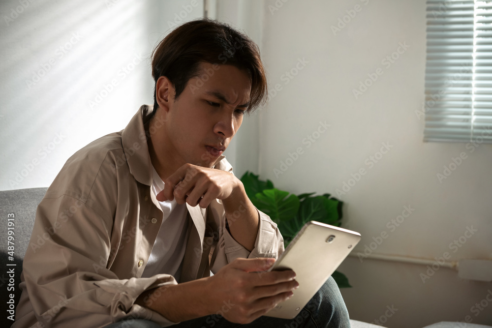 A tired and disappointing man sits on a sofa, surfing the Internet or shopping online via wireless internet on his tablet computer. While he was dissatisfied with the fact that working from home A tired and disappointing man sits on a sofa, surfing the Internet or shopping online via wireless internet on his tablet computer. While he was dissatisfied with the fact that working from home