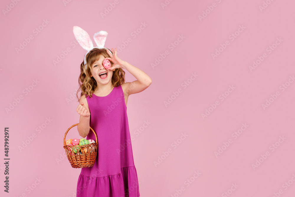 Happy child with long curly hair in fuchsia dress and bunny ears, smiling with open mouth and looking at camera while covering eye with Easter egg standing in pink studio with wicker basket