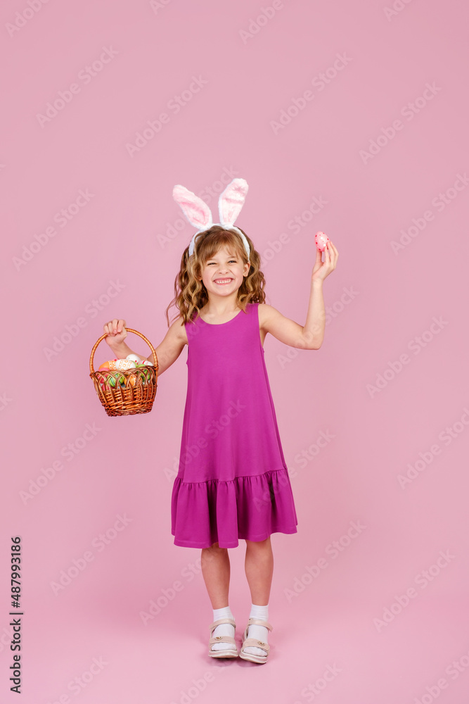 Adorable cute girl with blond curly hair in stylish dress and bunny ears headband, happily smiling and looking at camera while demonstrating wicker basket and colorful Easter egg, vertical, copy space