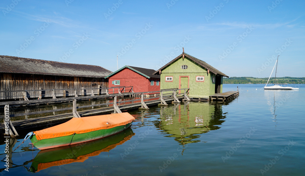 Fototapeta premium a long wooden pier leading to the colorful boat houses on serene sunlit lake Ammersee in the German fishing village Schondorf (Ammersee, Germany) 