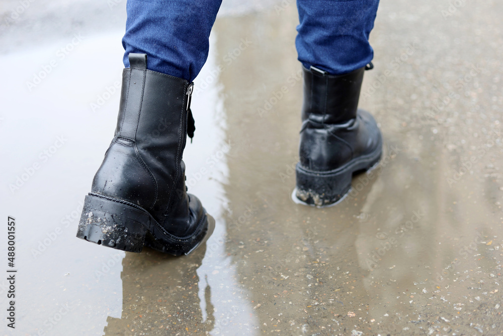 Female legs in black leather boots with reflection in puddle on a street. Rain in city, melting snow in spring, waterproof footwear Female legs in black leather boots with reflection in puddle on a street. Rain in city, melting snow in spring, waterproof footwear