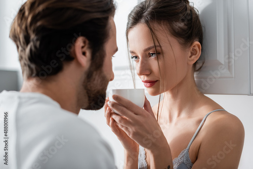young woman in bra holding cup and looking at blurred bearded boyfriend.