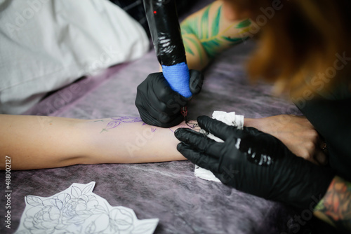 Tattoo parlor, pretty European red-haired girl makes a tattoo on arm. Woman creates a tattoo on a female hand by a professional tattoo artist
