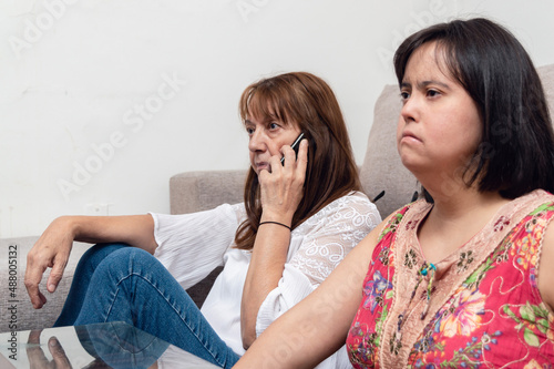 seated latin caucasian mother talking on the phone while her daughter watches television