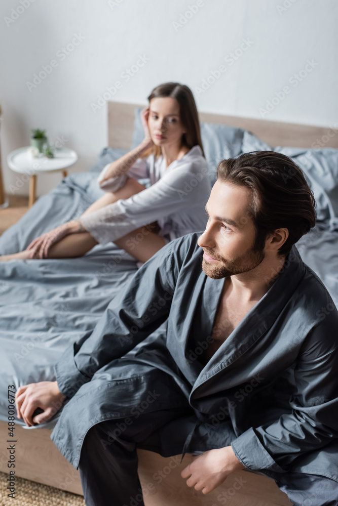 bearded man sitting on bed near blurred woman in silk robe. bearded man sitting on bed near blurred woman in silk robe.