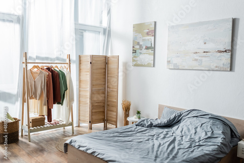 Φωτογραφία modern bed near folding screen and rack with clothes in bedroom.