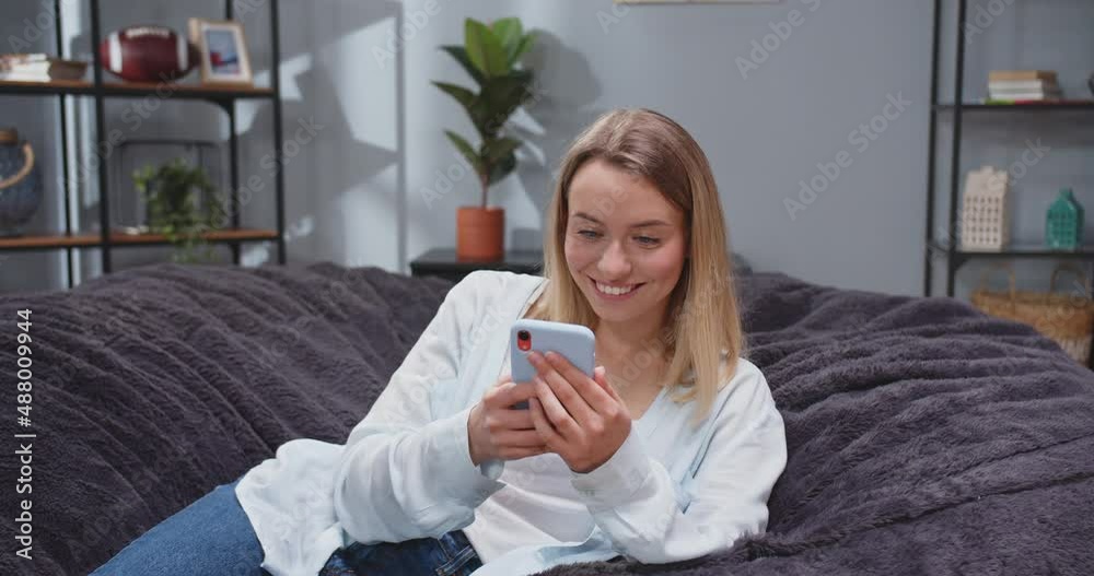 Close up front view shot of pretty caucasian woman reclining on sofa texting via phone. Female scrolling social media on gadget and smiling. People after work resting on sofa with smartphones. Close up front view shot of pretty caucasian woman reclining on sofa texting via phone. Female scrolling social media on gadget and smiling. People after work resting on sofa with smartphones.