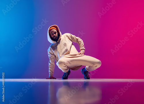 Studio shot of young flexible sportive man dancing breakdance in white outfit on gradient pink blue background. Concept of action, art, beauty, sport, youth