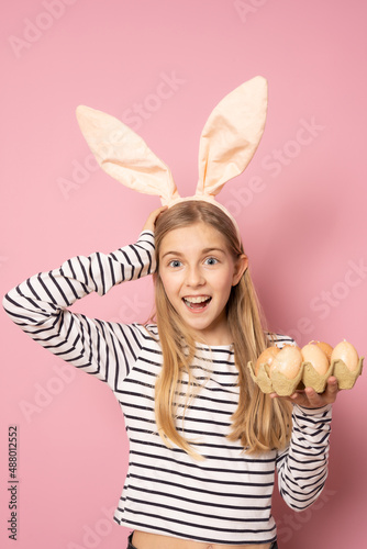 Happy Easter concept. Beautiful young girl smiling wearing rabbit ears holding Easter eggs on hand isolated over pink background.
