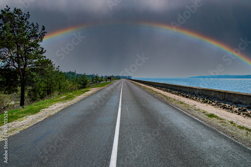 A rainbow with a thundercloud on the horizon of a highway. Rainbow in the storm. Asphalt pavement. Road markings on asphalt. Cloud horizon. Coastal road. Go on vacation. Summer season. © malaha