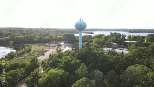 View of Excelsior, Minnesota, water tower and surrounding area including community garden and Lake Minnetonka in background. Road leading back into town that is tree lined. 