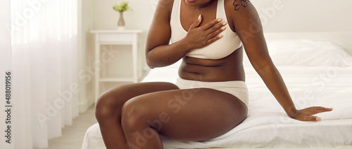 African American woman suffering from heart disease. Young black lady in white underwear suddenly feels pangs of pain, sits down on the edge of her bed and touches her chest. Cardiac pain concept photo