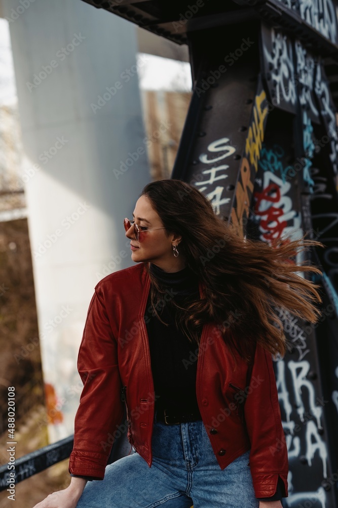 Berlin, Germany - 15.03.2020: Young female with long streaming dark hair wearing red leather jacket and red sunglasses. Graffiti on the background. 